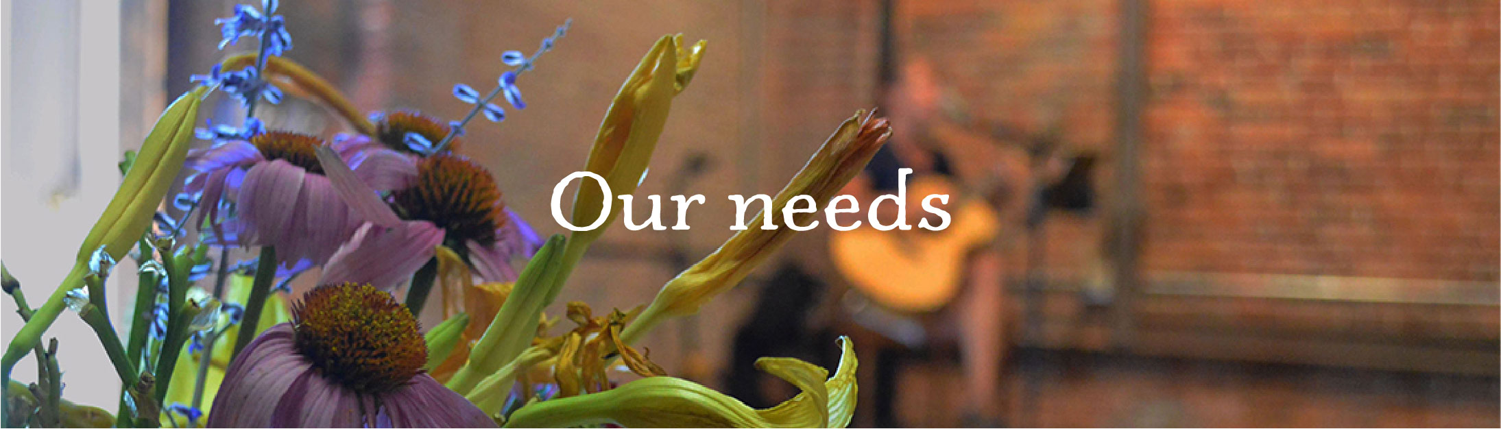 Flower bouquet closeup with blurred guitarist in front of brick wall, reads Our needs 